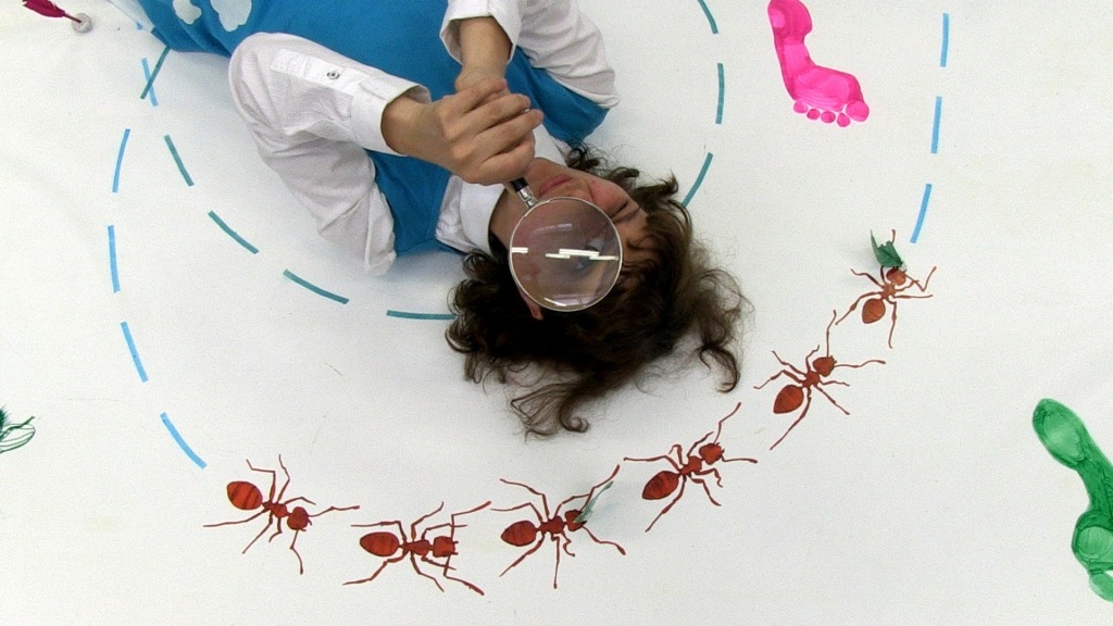 Deitada no chão e rodeada de desenhos de formigas, atriz Joana Providência olha através de uma lupa.