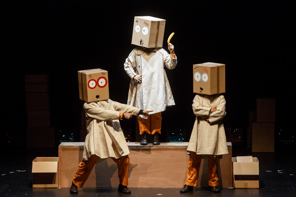 Três atores  da peça de teatro Frágil estão em cena com caixas de cartão sobre a cabeça.
