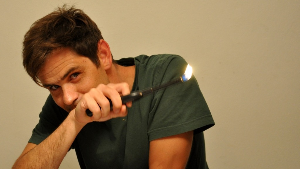 José Maria Vieira Mendes olha para a câmara. A mão esquerda que oculta a boca segura um longo isqueiro com a chama acesa na ponta.