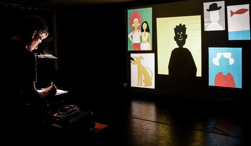 Através de um retroprojetor, João Fazenda projeta o desenho de um homem e uma mulher lado a lado, ambos apontando máquinas fotográficas no espetáculo Retrato Falado.
