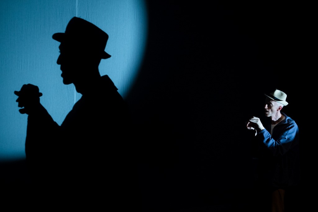 Fotografia de cena do espetáculo com um ator em contra-luz e uma grande sombra de fundo
