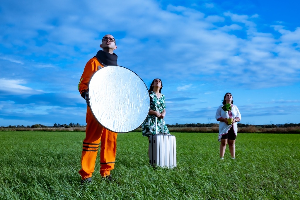 Três dos atores do Aldearã, no meio de um arrozal. Um está vestido de astronauta, uma agarra numa mala e a terceira uma planta.