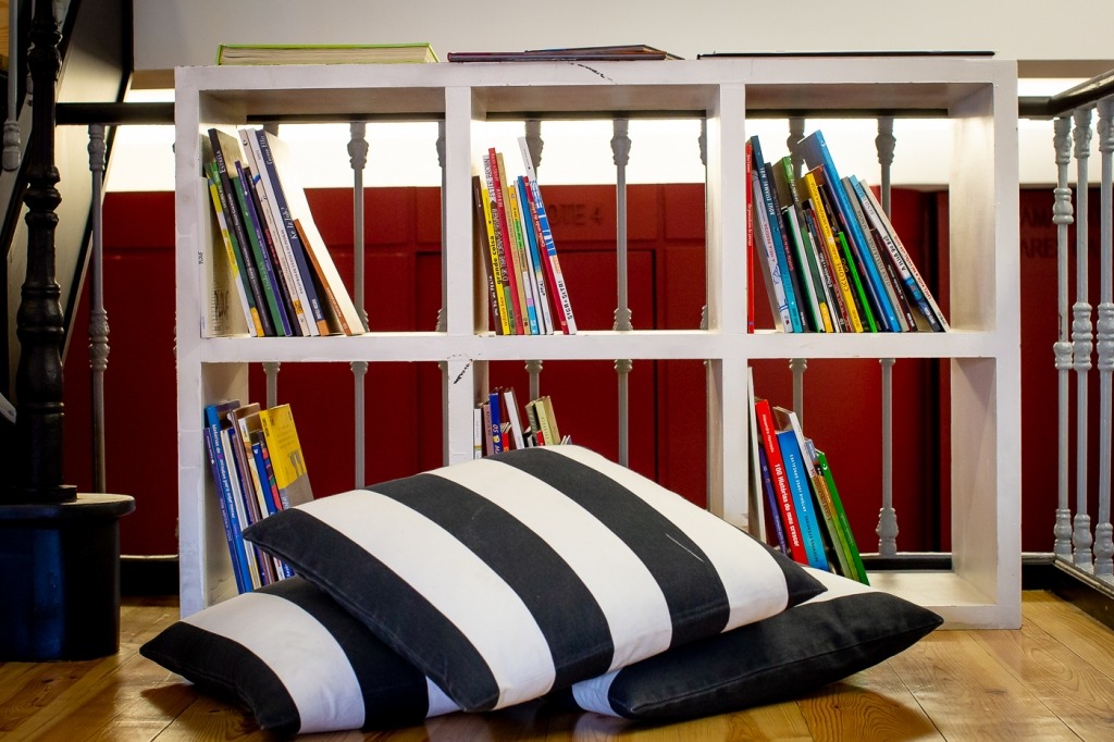 Uma estante branca com vários livros. No chão de madeira, em frente à estante, estão almofadas às riscas pretas e brancas.