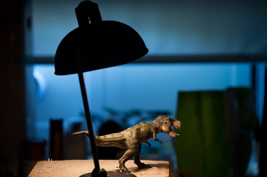 Figura de T-Rex pousada em cima de mesa junto a candeeiro de secretária