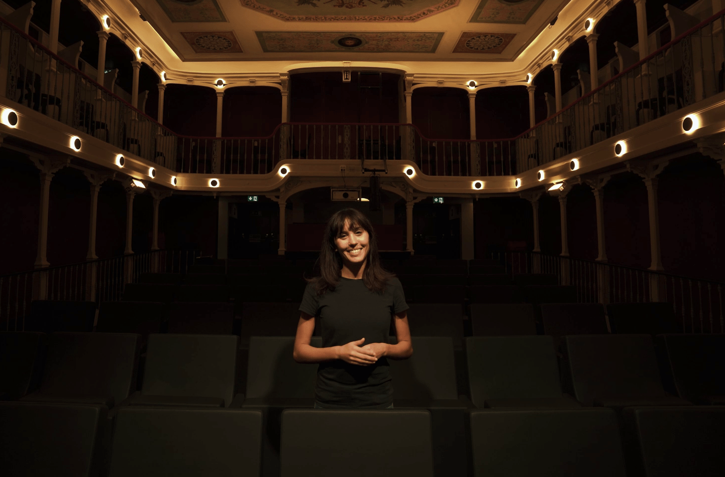Pessoa sorri no meio da plateia de um teatro.
