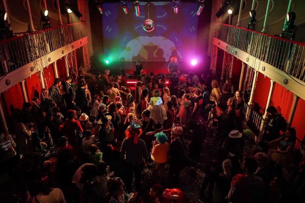 Pessoas mascaradas a dançar numa sala de teatro sem plateia e com bola de espelhos
