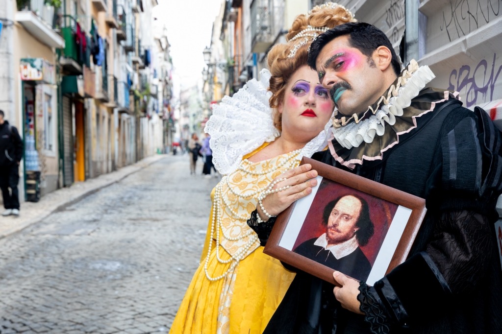 Um homem e uma mulher vestidos com vestuário da época Isabelina, com maquilhagem colorida nos rostos, estão encostados a uma parede grafitada. Nas mãos o homem segura uma moldura com um retrato do dramaturgo inglês William Shakespeare.