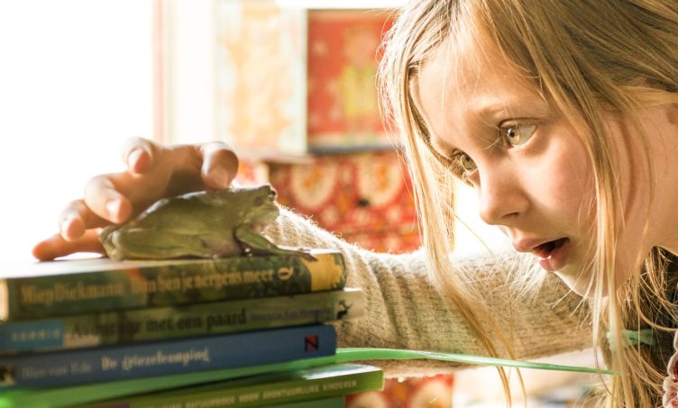 Uma rapariga olha e toca num sapo, pousado em cima de um monte de livros.