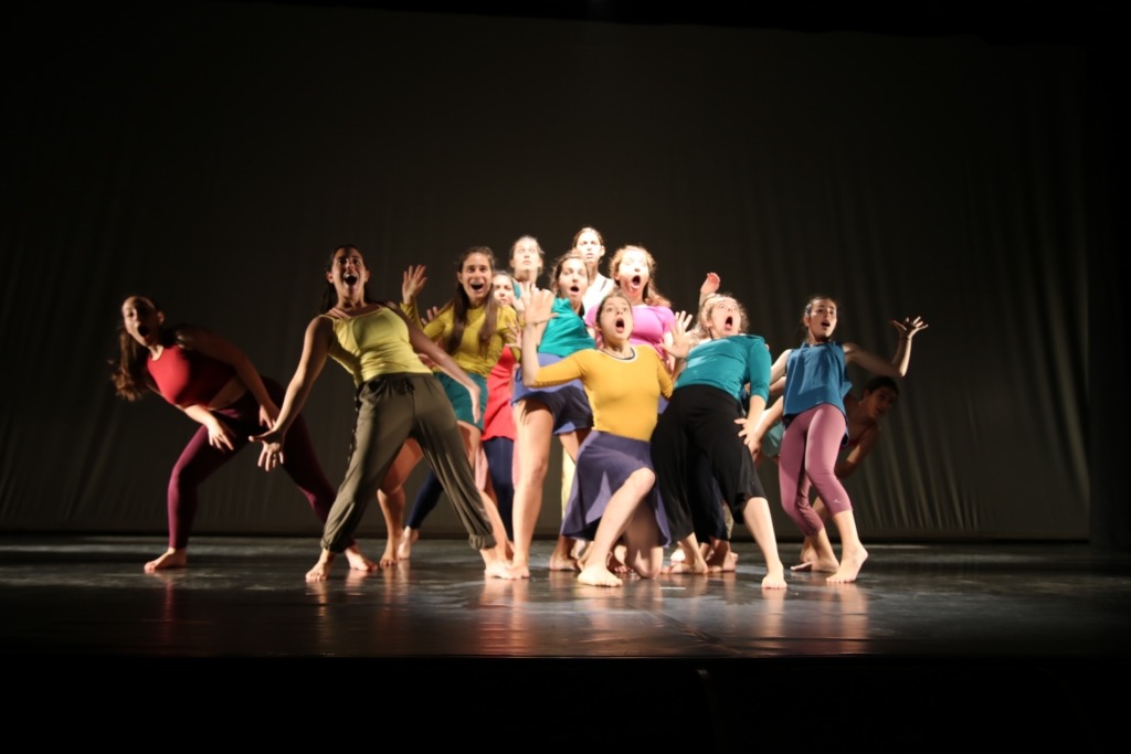 Grupo de mulheres em palco, com roupas coloridas. Estão de boca aberta, de semblante espantado