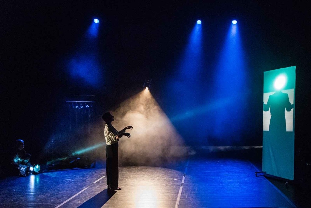 Em cima de um palco uma pessoa é iluminada por alguns projetores. À direita, a sombra da sua silhueta está projetada numa tela.