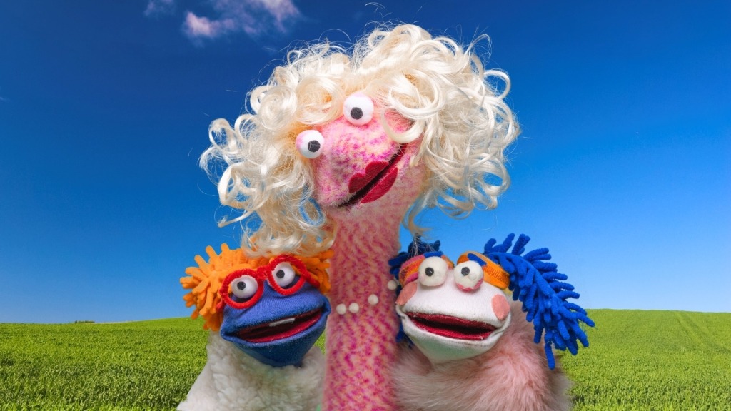 Três fantoches de meia com olhos grandes e cabelos coloridos olham diretamente para o observador. Atrás dos fantoches, encontra-se um prado verdejante e um céu azul.