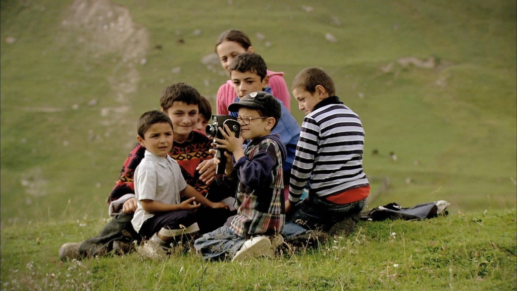 Um grupo de crianças reunidas em torno de um rapaz que segura uma câmara de filmar. O grupo está ao centro de uma paisagem verdejante.