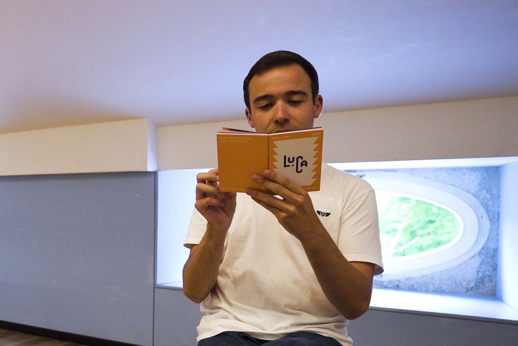 Homem de calças de ganga e t-shirt branca lê a brochura do LUCA. Este pequeno livro, de capa laranja e branca está erguido ao nível do rosto, tapando um pouco da sua face. 