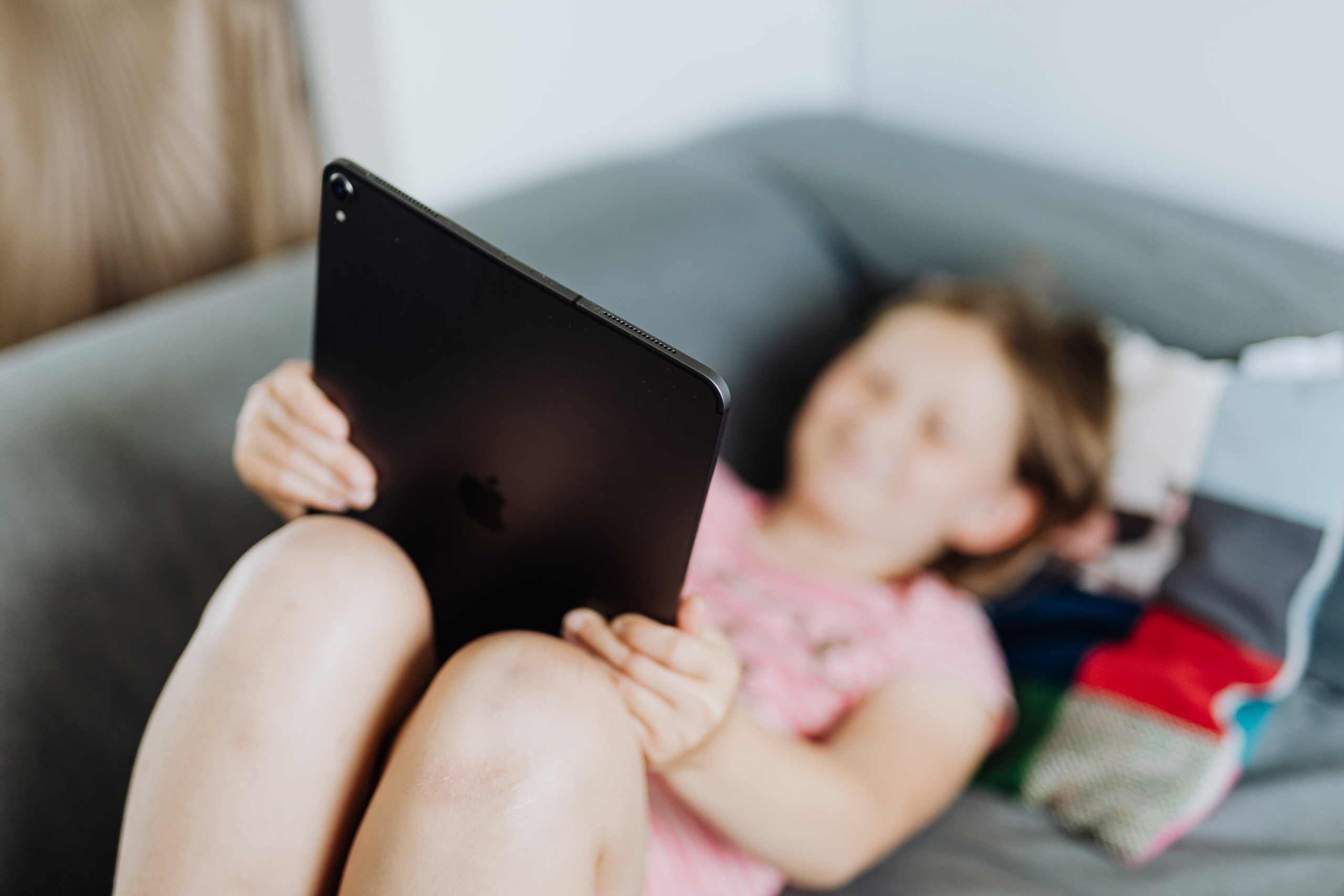 Criança, de rosto desfocado, deitada num sofá com um tablet apoiado nos joelhos.