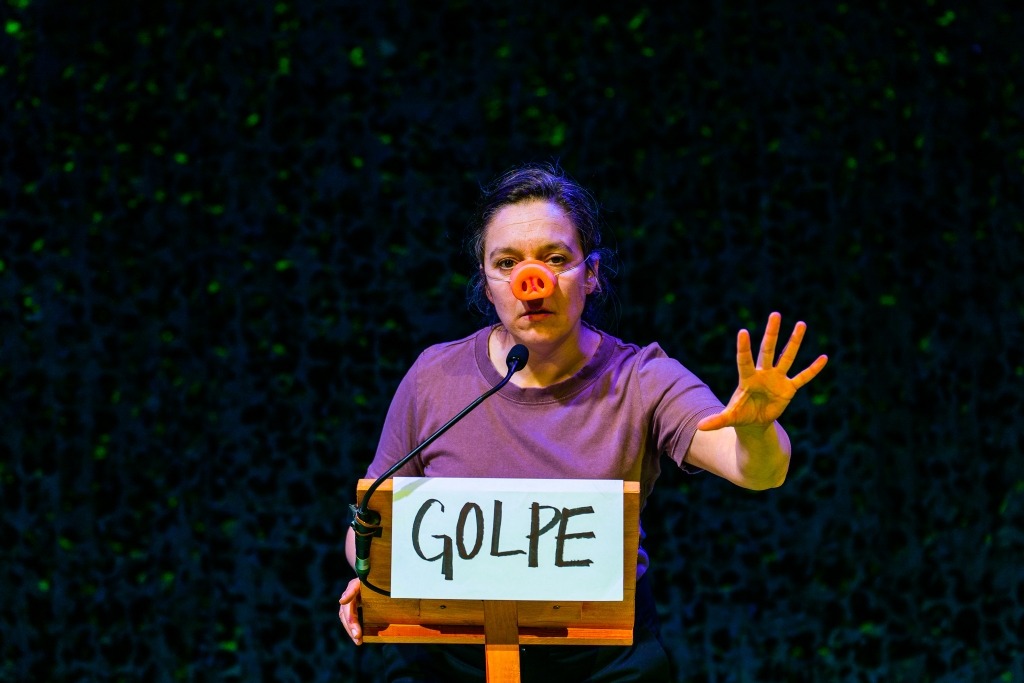 Atriz da cintura para cima atrás de um púlpito de discurso com a palavra Golpe escrita em letras maiúsculas. A atriz está com um nariz de porco e uma das mãos abertas erguida no ar.