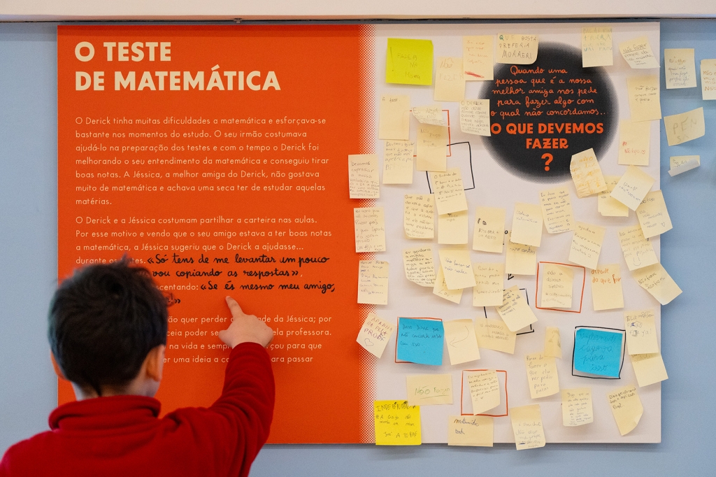 Criança de costas para a câmara aponta para um painel laranja com o título "O Teste da Matemática". O lado direito do painel está coberto de post-its.