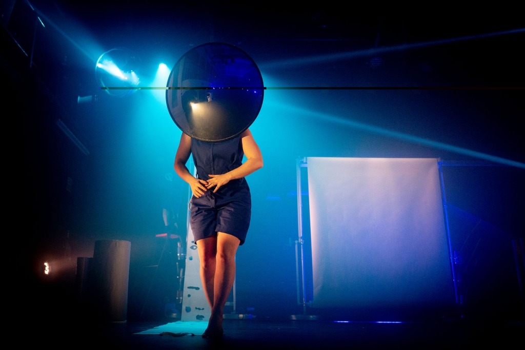 Num palco iluminado a azul, uma pessoa está de pé com grande espelho redondo à frente do rosto