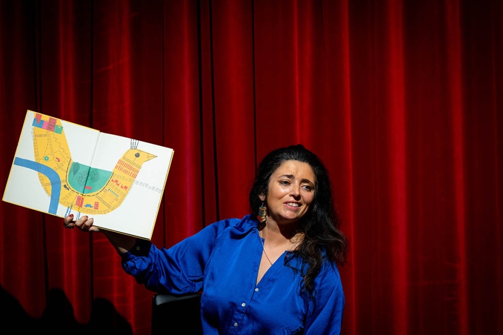 Mulher de camisa azul segura um livro ilustrado em frente de uma cortina vermelha