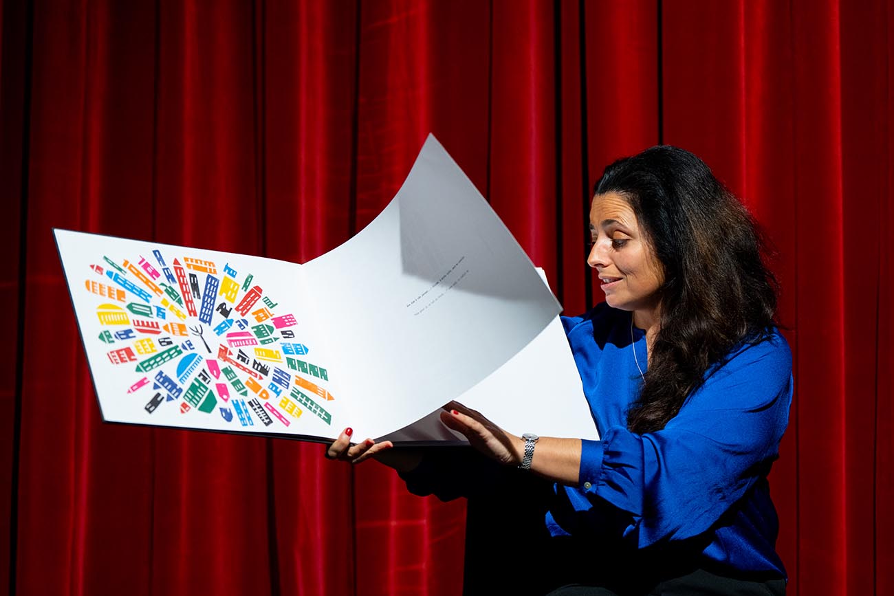 Bru Junça in profile and dressed in blue holds a white book with illustrations