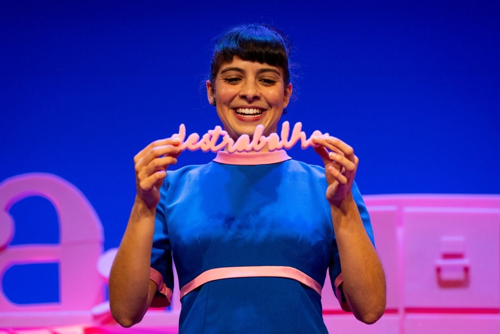 Atriz vestida de azul segura nas mãos uma palavra em esponja rosa: destrabalho.