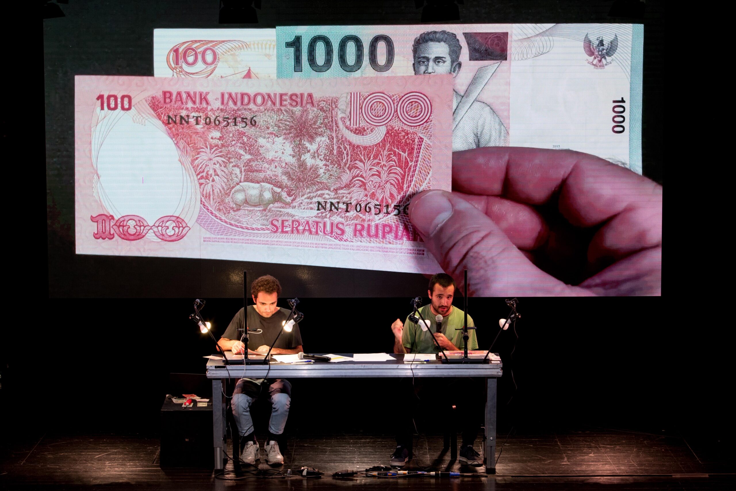Dois atores sentados numa mesa em palco. Atrás deles, uma tela exibe uma mão a manipular notas